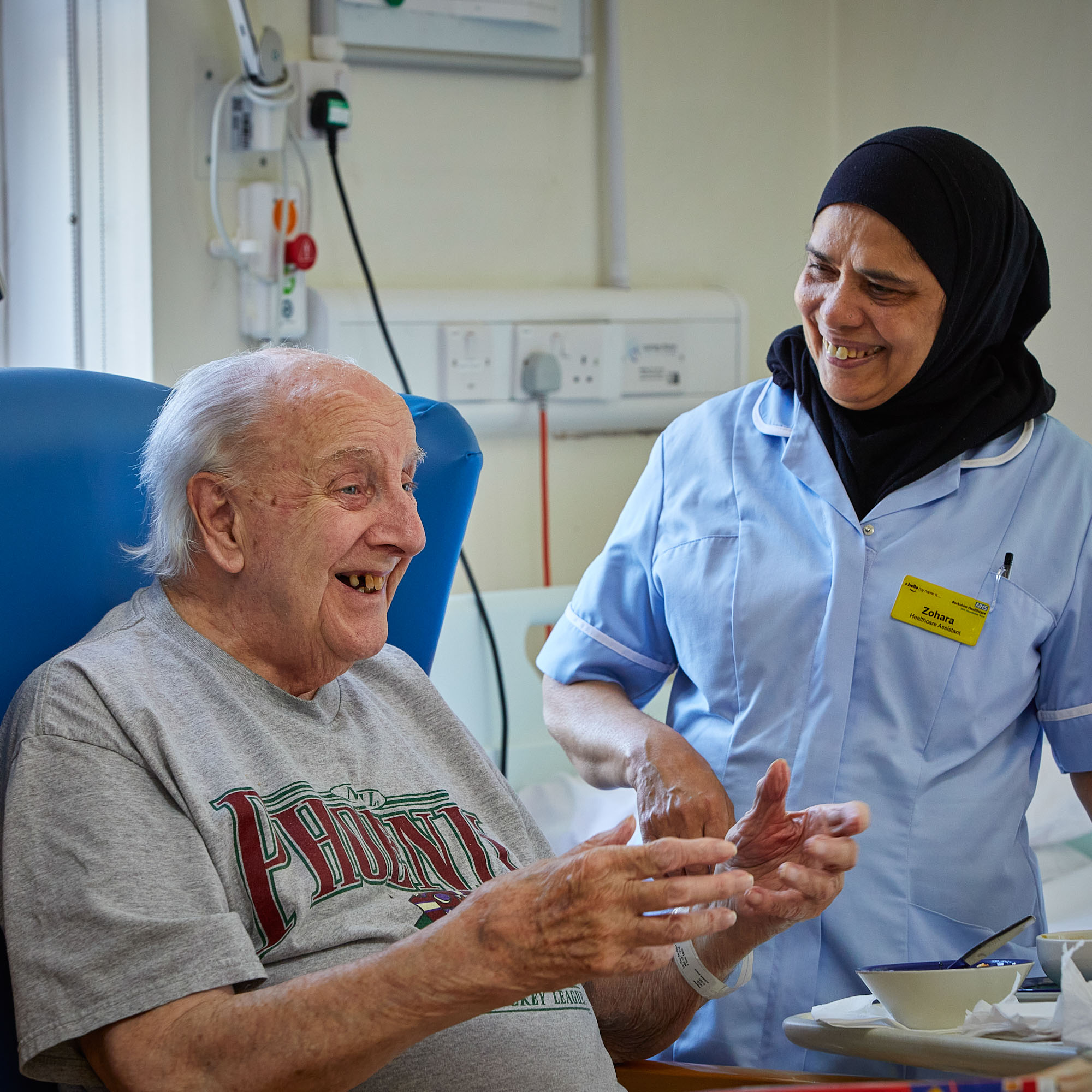 a healthcare assistant in a blue uniform smiling down at an elderly gentleman smiling with his hands in a open gesture sitting in a chair