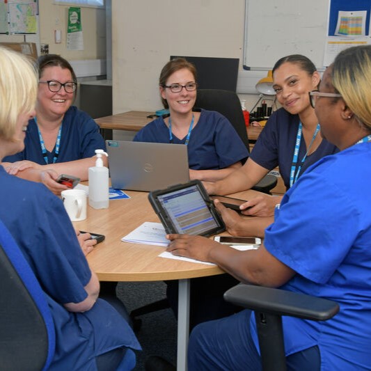 A group meeting between 5 nurses, sat around a table, with laptops and notepads.