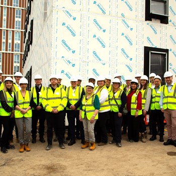 A group of people of different genders and race are stood in front of an unfinished building. They are wearing high vis vests and hard hats.