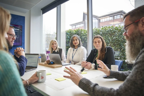 A group of 6 staff members, sitting around a table in a meeting