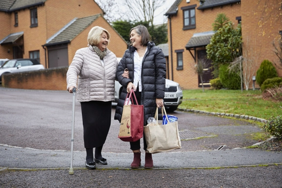 Woman with shopping bags helping woman with walking stick outside houses