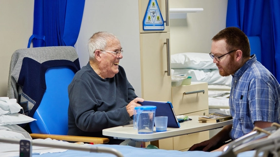 patient sitting by bed showing man something on his tablet