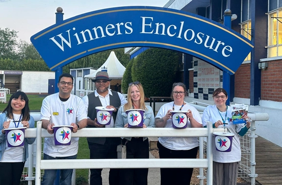 Fundraising team holding donation buckets under the sign for Ascot Racecourse's Winners Enclosure